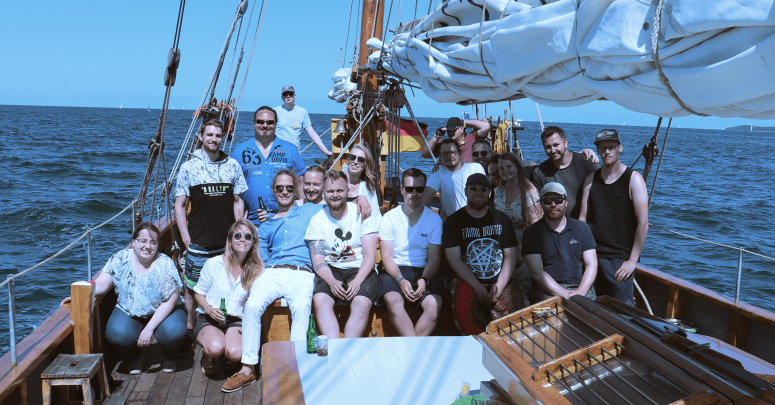 Das Team von Kontor Business IT um Co-Geschäftsführerin Carolina Wehrmann (hockend 2. v. l.) steht auf einem Segelschiff. Im Hintergrund ist das offene Meer zu erkennen.